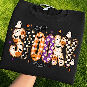 Pode incluir: Uma camisola preta de gola redonda com a palavra SPOOKY em letras grandes e estampadas. Cada letra tem um design único, incluindo bolinhas, xadrez laranja, flores roxas, fantasmas, abóboras, morcegos e estrelas espalhadas ao redor.