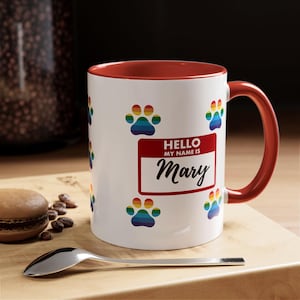 May include: White ceramic mug with a red interior and handle, decorated with rainbow paw prints. The mug features a name tag that says "Hello My Name Is Mary".