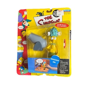 May include: A yellow Sideshow Mel action figure from The Simpsons in original packaging. The set includes a gray cannon accessory and a small red projectile. The yellow card backing features character illustrations and the text The Simpsons.