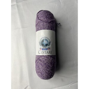 May include: A skein of heathered purple yarn with a white paper label wrapped around the center. The label features a circular logo with a blue cross and the text CESTARI printed in black capital letters below the emblem on the white background.