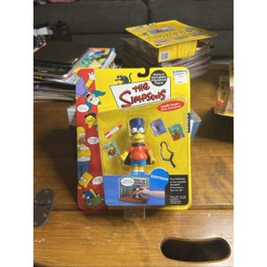May include: A Bartman action figure from The Simpsons World of Springfield series, packaged in a yellow blister card. The figure wears a red shirt, blue shorts, and a black cape, accompanied by a small rocket, a slingshot, and two miniature books.