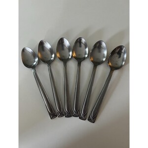 May include: A set of six silver-toned stainless steel spoons arranged in a fan pattern. Each spoon features a smooth, polished oval bowl and a handle decorated with a subtle, etched geometric line design near the base of the stem.
