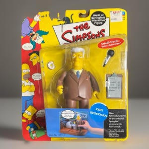 May include: A Kent Brockman action figure from The Simpsons World of Springfield series, packaged in a yellow blister card. The figure wears a brown suit and blue tie, accompanied by a microphone and a clipboard. The packaging features character graphics.