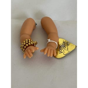 May include: Two doll arms with a light skin tone. One arm has a gold beaded bracelet. The other arm has a white string tied around the wrist. A gold heart-shaped tag with the words "Vogue Dolls" is also present.