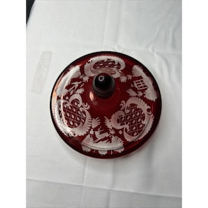 May include: A circular ruby red glass lid featuring intricate white etched patterns of stylized foliage, geometric lattice shapes, and a small deer figure. The lid has a smooth, rounded dark red glass knob handle centered on the top surface.