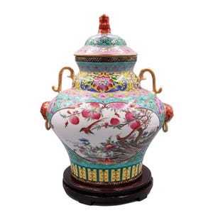 May include: A decorative ceramic vase with a lid, featuring a detailed floral and bird design in shades of pink, green, yellow, and red. The vase has gold handles and rests on a dark brown wooden base. The vase is approximately 14 inches tall.