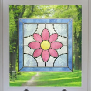 Puede incluir: Un panel de ventana de vidriera cuadrada con una flor rosa y un centro amarillo. La flor se encuentra sobre un fondo blanco y azul claro, enmarcada por un borde azul. La ventana está en un marco blanco, con un fondo verde lleno de árboles.