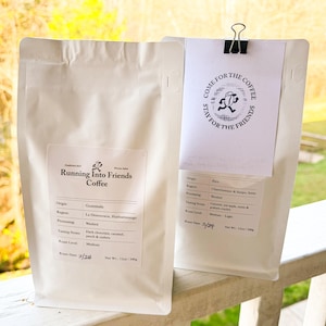 May include: Two white coffee bags with labels. One bag reads "Running Into Friends Coffee" with details about the origin, region, processing, tasting notes, and roast level. The other bag has a paper clipped on it that says "Come for the coffee, stay for the friends."