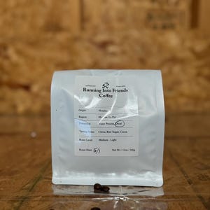 May include: A white coffee bag with the text "Running Into Friends Coffee" and details about the coffee's origin, processing, and tasting notes. The bag contains 12oz of coffee. Several coffee beans are scattered in front of the bag.