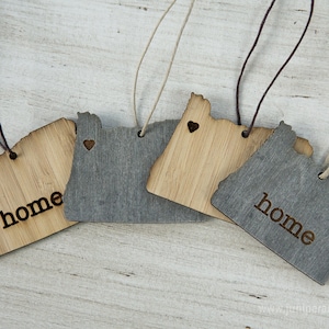 May include: Four wooden ornaments shaped like the state of Oregon. Two are natural wood with the word "home" engraved on them. Two are painted gray with a small heart cutout and the word "home" engraved on them.