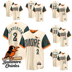 Op de afbeelding: Crèmekleurige honkbalshirts met groene en oranje accenten. De shirts tonen het woord "BMORE" en spelersnamen en -nummers. Het logo van de Baltimore Orioles is zichtbaar. De shirts hebben korte mouwen.