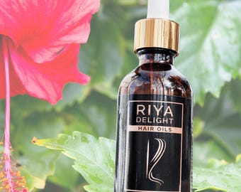 May include: A brown glass bottle with a gold lid and a dropper top. The bottle is labeled "RIYA DELIGHT HAIR OILS" and contains 2 fl oz (60 ml) of hair oil. The bottle is sitting on a green leafy background.