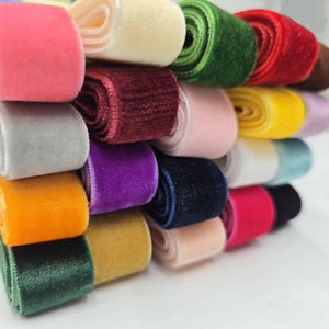 Puede incluir: Una colección de rollos de cinta de terciopelo en varios colores, incluyendo rosa, crema, verde, rojo, burdeos, rosa claro, amarillo, gris, morado, azul marino, naranja, mostaza y negro, dispuestos en una pila para mostrar la textura.