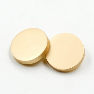 Puede incluir: Dos objetos circulares de color dorado con un acabado mate. Los objetos están colocados cerca uno del otro, solapándose ligeramente. La iluminación resalta los bordes lisos y redondeados y las sutiles variaciones del tono dorado.