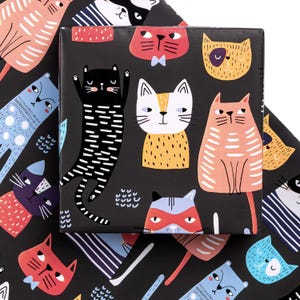 Peut inclure: Papier cadeau noir avec un motif répété de visages de chats de dessins animés colorés. Les chats ont diverses expressions et sont dans des tons de rouge, orange, bleu et blanc. Le papier est plié sur un paquet carré.