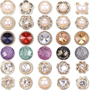 Puede incluir: Una colección de botones decorativos en varios diseños y colores. Los botones presentan perlas, cristales y detalles metálicos. Los estilos incluyen formas florales, redondas y geométricas, con una gama de colores que van desde el dorado hasta el iridiscente.