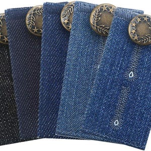 May include: Five denim fabric extenders in various shades of blue and black. Each extender features a decorative bronze button with a leaf and star design. The extenders have a buttonhole for attachment to pants.