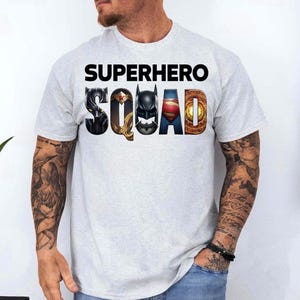 Puede incluir: Camiseta gris claro con las palabras "SUPERHERO SQUAD" en negro. La palabra "SQUAD" presenta logotipos de superh&eacute;roes. La camiseta es de manga corta y la lleva una persona con tatuajes en los brazos.