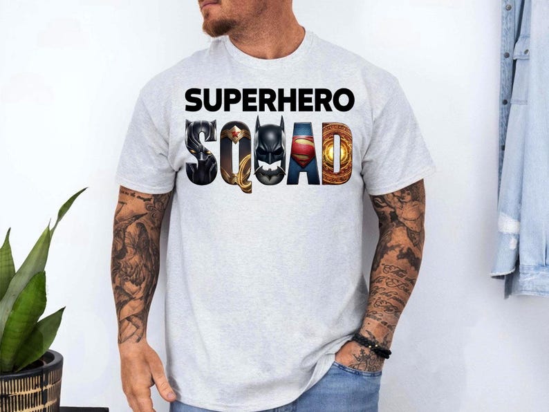 Puede incluir: Camiseta gris claro con las palabras "SUPERHERO SQUAD" en negro. La palabra "SQUAD" presenta logotipos de superh&eacute;roes. La camiseta es de manga corta y la lleva una persona con tatuajes en los brazos.