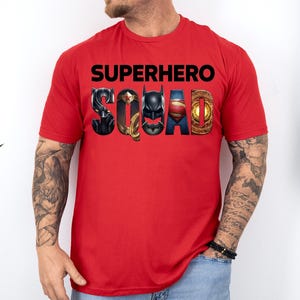 Puede incluir: Una camiseta roja con la frase "SUPERHERO SQUAD" en letras negras. La palabra "SQUAD" est&aacute; dise&ntilde;ada con logotipos de superh&eacute;roes, incluyendo Pantera Negra, Wonder Woman, Batman, Superman y un emblema dorado.