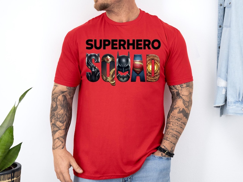 Puede incluir: Una camiseta roja con la frase "SUPERHERO SQUAD" en letras negras. La palabra "SQUAD" est&aacute; dise&ntilde;ada con logotipos de superh&eacute;roes, incluyendo Pantera Negra, Wonder Woman, Batman, Superman y un emblema dorado.