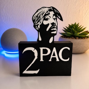 Peut inclure: Une enseigne décorative noire et blanche représentant un portrait stylisé de Tupac Shakur au-dessus du texte 2PAC. Le design est réalisé dans un style graphique à fort contraste avec une base noire solide et des lettres blanches.