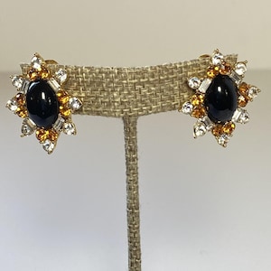 May include: A pair of gold-toned clip-on earrings. Each earring features a large, oval, black center stone surrounded by a starburst design of clear, amber, and white crystals. The earrings are displayed on a neutral-toned, textured surface.
