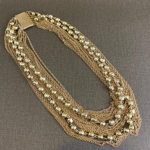 Puede incluir: Collar dorado con múltiples hilos de cadenas delicadas y pequeñas cuentas redondas. El collar tiene un cierre rectangular y se muestra sobre una superficie gris neutra. El diseño en capas añade textura e interés visual a esta pieza de joyería.