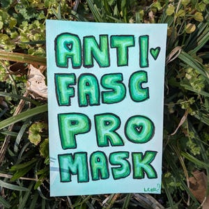 May include: A hand-painted watercolor sign with the words "ANTI FASC PRO MASK" in green block letters on a light blue background. The letters are outlined in black and have a heart symbol in the letters A, C, and O. The sign is resting on a bed of green grass.