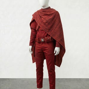 May include: A red costume on a mannequin. The outfit includes a long-sleeved shirt, pants, a cape, and a belt with pouches. The costume is a solid red color, with a textured appearance. The mannequin has a white head and hands.