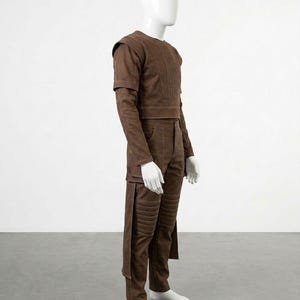 Pode incluir: Um conjunto marrom de duas pe&ccedil;as em um manequim. A parte superior tem mangas compridas e ombreiras. As cal&ccedil;as t&ecirc;m acolchoamento nos joelhos e uma aba lateral. O conjunto &eacute; feito de um material semelhante a camur&ccedil;a.