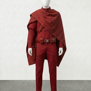 May include: A complete red costume displayed on a mannequin. The outfit includes a long-sleeved shirt, pants, a cape, and a belt with pouches. The costume is made of a textured fabric.