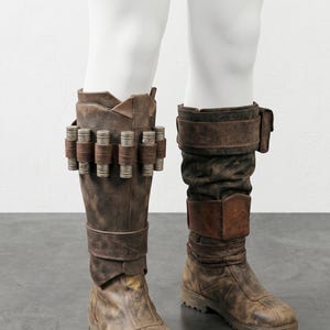 K&ouml;nnte beinhalten: Braune Lederstiefel mit Used-Look, verziert mit dekorativen Metallzylindern und Riemen. Die kniehohen Stiefel haben ein robustes, abgenutztes Aussehen, was auf ein Vintage- oder Kost&uuml;mdesign hindeutet.