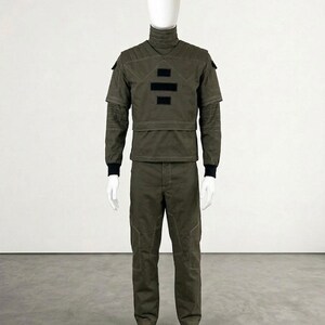 May include: Olive green military-style uniform displayed on a mannequin. The outfit includes a high-collared jacket with black accents, matching pants, and black socks. The jacket features a unique design element.