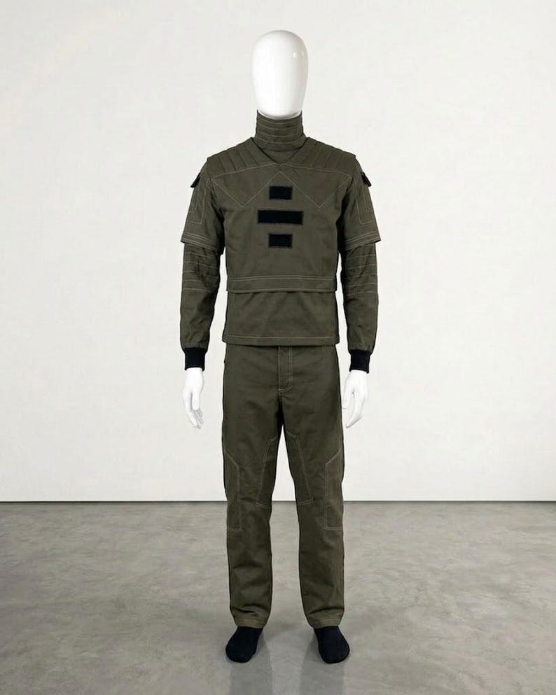 May include: Olive green military-style uniform displayed on a mannequin. The outfit includes a high-collared jacket with black accents, matching pants, and black socks. The jacket features a unique design element.