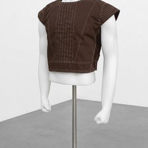 Pode incluir: Uma parte superior marrom de manga curta com detalhes de costura verticais e decote redondo. A parte superior &eacute; exibida em um torso de manequim branco com bra&ccedil;os, montado em um suporte de metal prateado. O fundo &eacute; uma parede branca lisa e um ch&atilde;o cinza.