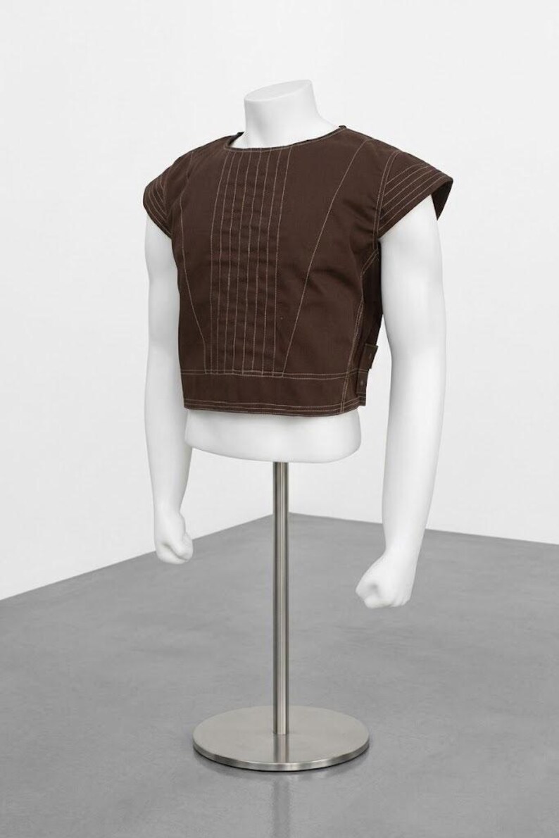 Pode incluir: Uma parte superior marrom de manga curta com detalhes de costura verticais e decote redondo. A parte superior &eacute; exibida em um torso de manequim branco com bra&ccedil;os, montado em um suporte de metal prateado. O fundo &eacute; uma parede branca lisa e um ch&atilde;o cinza.