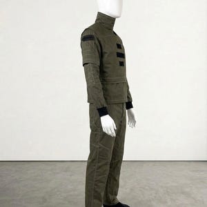 May include: An olive green jumpsuit with a high collar, black accents, and a structured design. The outfit features a long-sleeved top with patches and a matching pair of pants. The mannequin is wearing black socks.