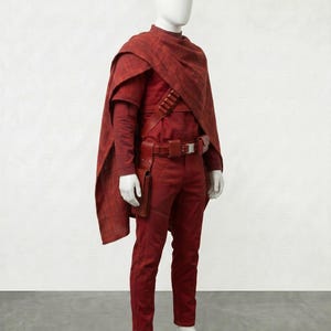 May include: A complete red costume on a mannequin. The outfit includes a long-sleeved shirt, pants, a cape, and a belt with a holster. The costume is made of a textured fabric and is displayed against a plain white background.