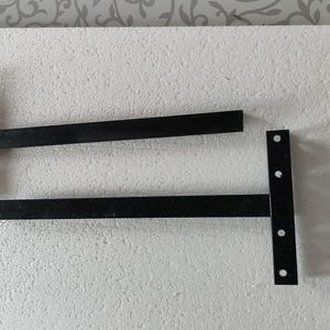 Puede incluir: Dos soportes de estante de metal negro, cada uno con una placa de montaje vertical con tres orificios circulares preperforados y un brazo de soporte horizontal que se extiende hacia afuera, diseñados para instalaciones de estantes.