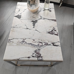 Puede incluir: Mesa de centro rectangular con tapa de mármol blanco con vetas grises oscuras y burdeos. La mesa tiene una estructura de metal pulido. Un cuenco decorativo y una botella de cristal están encima.