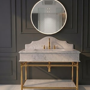 May include: A white marble bathroom vanity with a gold metal frame and a matching marble shelf. The vanity features a gold faucet and two handles, topped with a curved marble backsplash. Above sits a round, wall-mounted mirror with integrated lighting.