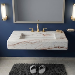 May include: A modern bathroom with a marble sink, gold fixtures, and a navy blue wall. The sink has a rectangular basin and is mounted on the wall. Two gold sconces flank a mirror above the sink. A towel rack with colorful towels is on the left.