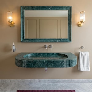 May include: A bathroom with a green marble sink and matching framed mirror. The sink is mounted on the wall and has a built-in drain. Two sconces with glass shades flank the mirror. A white towel hangs on a chrome towel ring.