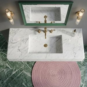 May include: A modern bathroom vanity with a white marble countertop and integrated sink. The vanity features gold-toned fixtures, a green-framed mirror, and two wall sconces. A pink circular rug sits on the floor, with green marble flooring and a white towel.