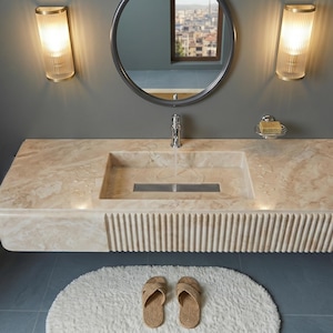 May include: A modern bathroom scene featuring a beige marble sink with a rectangular basin and a fluted front. Two sconces flank a round mirror above the sink. A chrome towel rack holds towels, and a white rug with sandals sits on the floor.