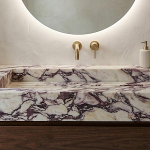 May include: A luxurious bathroom vanity with a marble countertop featuring a purple and white veined pattern. The vanity includes a built-in sink, gold fixtures, and a matching soap dispenser and tumbler. A round mirror with a soft glow is above the vanity.