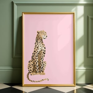 May include: A framed art print featuring a seated leopard illustration. The leopard has a tan coat with black spots, set against a soft pink background. The artwork is enclosed in a gold frame, adding a touch of elegance to the piece.
