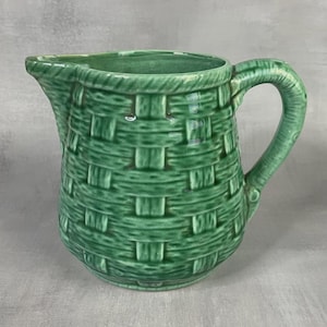 Puede incluir: Una jarra de cerámica verde con una textura en relieve que imita el tejido de una cesta en toda su superficie exterior. El recipiente incluye un asa curva en el lado derecho y un pico vertedor prominente en el borde superior izquierdo, esmaltado.
