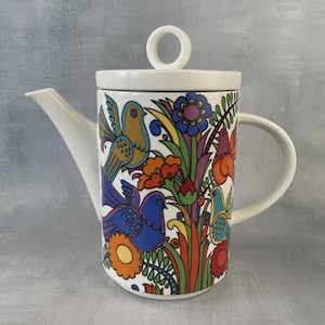 Puede incluir: Una tetera de cerámica blanca con un colorido patrón gráfico de aves, flores y follaje estilizados en tonos azul, naranja, amarillo, morado y verde. La tetera tiene un cuerpo cilíndrico, un asa lateral, un pico vertedor y una tapa con un asa circular.